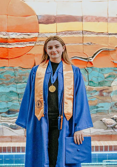Person stands  in cap and gown in front of sunset mural