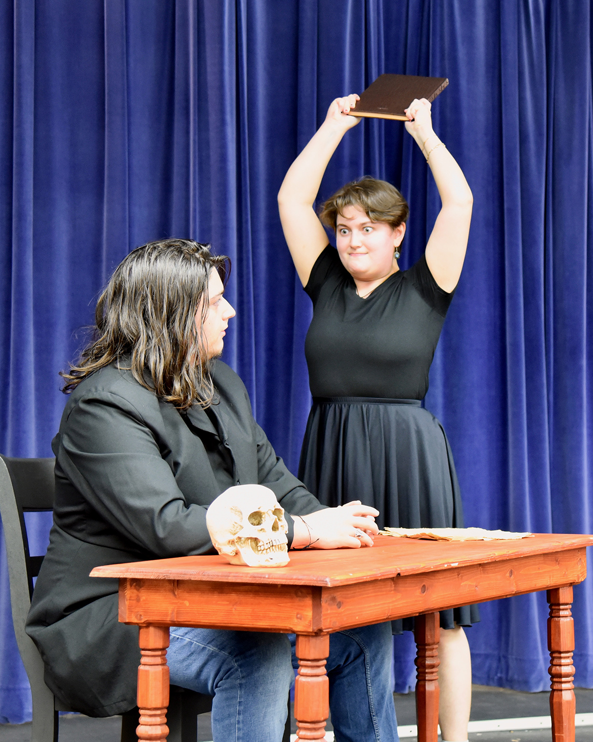 A person holds a book above their head while looking at another person sitting at a table with a skull on it.
