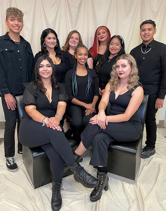 9 cosmetology students pose in all black in front of white backdrop.