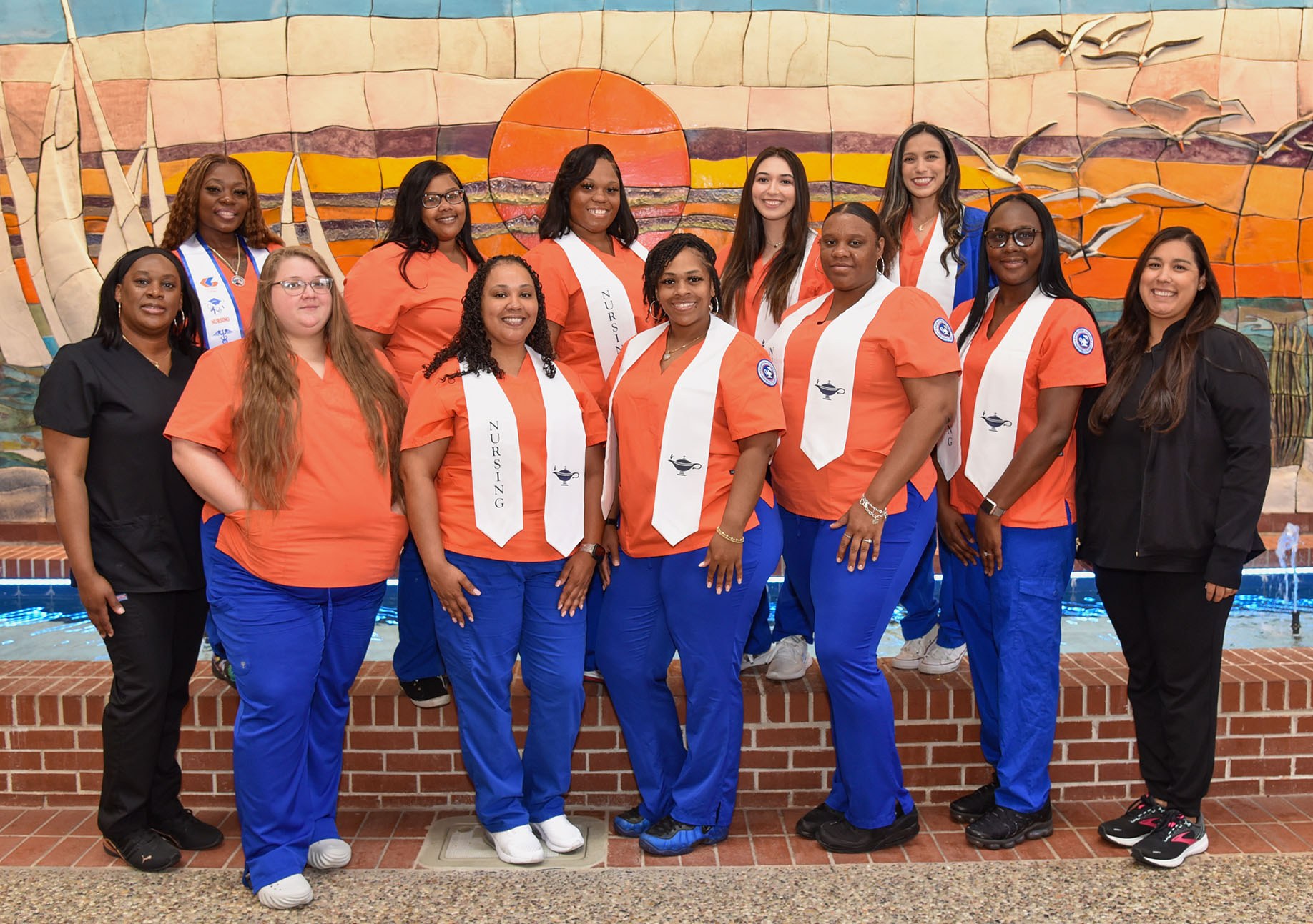 12 nursing students in scrubs pose in front of a sunset mural